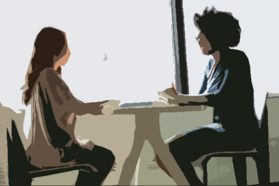 Blurred image of two women talking at a table in an office setting.